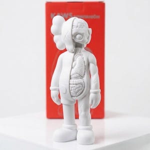 May include: A white vinyl figure with exposed internal organs on one side. The figure has a signature X-shaped eye design and a rounded head. A red box with text is in the background.