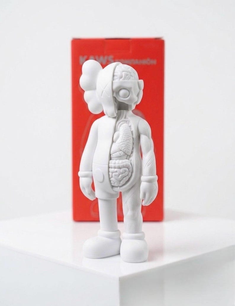 May include: A white vinyl figure with exposed internal organs on one side. The figure has a signature X-shaped eye design and a rounded head. A red box with text is in the background.