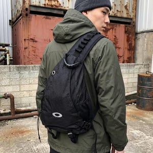 May include: A black sling backpack featuring a single wide shoulder strap with gear loops, a top handle, a side mesh pocket, and a white Oakley logo on the lower front. The bag is constructed from a textured fabric and includes a silver metal eyelet.