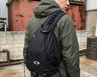 Puede incluir: Una mochila bandolera negra con una sola correa ancha para el hombro con trabillas, un asa superior, un bolsillo lateral de malla y un logotipo blanco de Oakley en la parte inferior. La bolsa es de tela texturizada e incluye un ojal metálico.