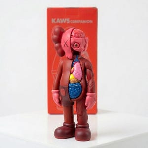 May include: A maroon KAWS Companion figure with exposed internal organs, including a brain, lungs, and intestines. The figure has a partially dissected appearance, with one side revealing the internal anatomy. The figure is standing in front of a red box with the text "KAWS COMPANION".