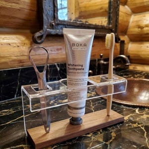 May include: A clear acrylic and wood toothbrush holder with a tube of Boka whitening toothpaste, a toothbrush, and a tongue scraper. The toothpaste tube is silver with the words "Bright White" and "Whitening Toothpaste" printed on it.