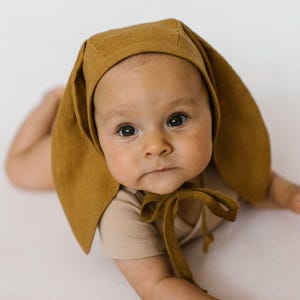 May include: A mustard yellow bonnet featuring long, pointed rabbit-style ear flaps that extend down the sides of the face. The headwear is constructed from a textured fabric and includes matching fabric ties that fasten securely under the chin.