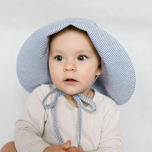 Può includere: Un cappello da sole a tesa larga con un motivo a righe sottili blu e bianche, dotato di lacci in tessuto coordinati che si allacciano sotto il mento. Il cappello è indossato da un bambino piccolo con una maglia a maniche lunghe a coste.