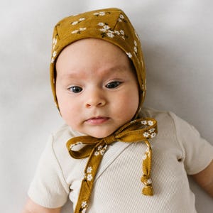 Peut inclure: Un bonnet jaune moutarde orné d'un petit motif floral blanc avec un lien à nouer sous le menton, porté avec un body à manches courtes côtelé de couleur crème. Le bonnet présente une couture structurée et des liens en tissu souple.