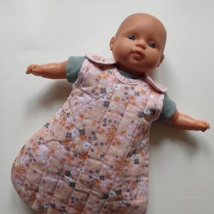 Könnte beinhalten: Eine Babypuppe in einem rosa gesteppten Schlafsack mit Blumenmuster in Braun-, Weiß- und Grautönen. Die Puppe hat einen kahlen Kopf und trägt ein grünes Oberteil. Der Schlafsack hat ein gestepptes Design.