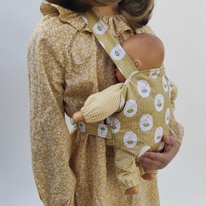 May include: A quilted doll carrier featuring a mustard yellow fabric with a white circular pattern containing a small green and pink flower motif. The carrier has matching mustard yellow straps and is worn over a mustard yellow floral dress.