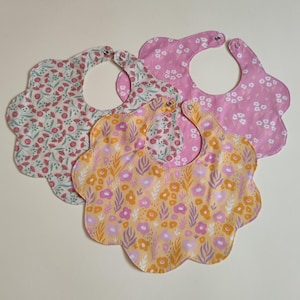 Può includere: Tre bavaglini con bordo smerlato e motivi floreali. Un bavaglino ha uno sfondo bianco con piccoli fiori rossi e verdi, uno è rosa con fiori bianchi e uno è giallo con motivi di fiori e foglie viola e arancioni. Ognuno ha una chiusura a scatto.