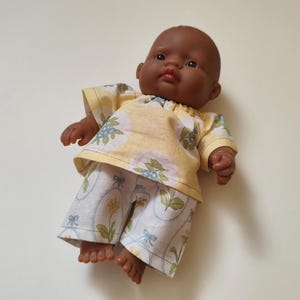 Op de afbeelding: Een donkerhuidige babypop draagt een geel shirt met korte mouwen met een blauw en groen bloemenpatroon en een bijpassende witte broek met een botanische print in ovale kaders met blauwe strikjes. De pop heeft een glad hoofd en bruine ogen.
