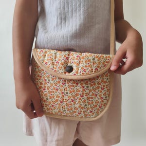 Può includere: Una borsa a tracolla trapuntata con un motivo floreale su piccola scala nei toni dell'arancione, giallo e verde su sfondo crema. La borsa presenta una patta arrotondata, una chiusura con bottone marrone scuro e bordi beige chiaro.
