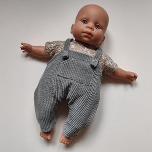 Puede incluir: Muñeca bebé con ojos azules, vestida con una camisa con estampado floral y un peto de pana gris. La muñeca está tumbada sobre una superficie blanca con los brazos extendidos.