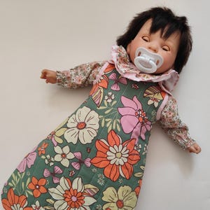 May include: A baby doll with dark hair and a pacifier, wearing a floral print sleep sack and a matching floral top with a ruffled collar. The sleep sack is green with large orange, pink, and white flowers.