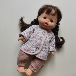Puede incluir: Una muñeca con cabello castaño trenzado, vestida con una chaqueta con estampado floral con ribete rosa y pantalones a juego. La muñeca tiene tez clara y está sobre una superficie blanca.