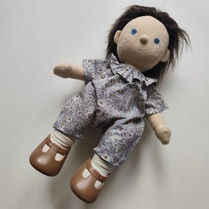 May include: A handmade doll with a fabric body, brown hair, and blue eyes. The doll wears a floral print jumpsuit and brown shoes. The doll's face is hand-stitched with simple features. The doll is lying down.