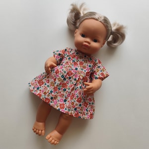 May include: A doll with blonde pigtails wearing a floral print dress in shades of red, pink, blue, and green. The doll has a fair complexion and is lying on a white surface.