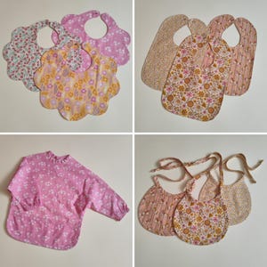 May include: A collection of baby accessories including three scalloped-edge bibs with floral patterns, three elongated bibs with floral prints, a long-sleeved pink smock with white flowers, and three rounded bibs with tie closures and floral designs.