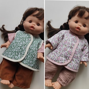 Op de afbeelding: Een pop met bruin haar in vlechten, gekleed in een gewatteerd vest met bloemenpatroon, een shirt met korte mouwen en een broek. De linkerpop draagt een groen vest en bruine broek, de rechterpop een wit vest met bloemen en een roze broek.