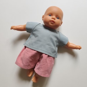 May include: A doll dressed in a light blue t-shirt and pink shorts. The doll has a bald head, blue eyes, and outstretched arms. The doll's outfit is simple and casual.