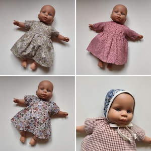 May include: Four baby dolls dressed in floral print dresses and a bonnet. The dresses are in shades of pink, brown, and blue, with various floral patterns. The dolls have blue eyes and are lying on a white surface.