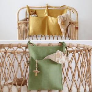 May include: A fabric storage organizer with two pockets, available in mustard yellow or sage green, featuring tie-top straps for attachment to a crib. The pockets contain a small plush animal, a light-colored cloth, and a pacifier on a braided cord.
