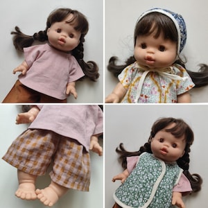 Könnte beinhalten: Eine Vinylpuppe mit braunem Haar in zwei Zöpfen, bekleidet mit verschiedenen Outfits, darunter ein altrosa Oberteil, braune karierte Shorts, eine geblümte Haube, ein Oberteil mit Vogelmuster und eine grüne Blumenweste mit weißem Saum und drei Knöpfen.