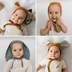 May include: A collection of four baby bonnets in various colours and styles, including a rust-coloured bonnet with small ears, a plain orange bonnet, a slate blue bonnet with long rabbit-like ears, and a mustard yellow bonnet with flower-shaped ears.