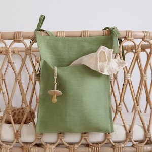 May include: A sage green linen crib organizer with two pockets and tie closures. A pacifier and a white muslin cloth are in the pockets. The organizer is attached to a woven rattan crib.