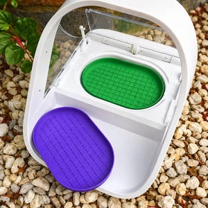 Puede incluir: Un comedero automático para mascotas de plástico blanco con tapa transparente abatible, que incluye una bandeja ovalada verde extraíble y otra bandeja ovalada morada, ambas con un patrón de rejilla y acabado en blanco liso.