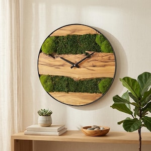 May include: A circular wall clock featuring a natural wood grain face with horizontal bands of vibrant green preserved moss. The clock has a thin black metal frame and simple black hour and minute hands centered on the wood surface.