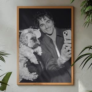 Puede incluir: Fotografía enmarcada en blanco y negro de una persona sosteniendo un perro pequeño mientras se toma una selfie con un teléfono inteligente. La fotografía se muestra en una pared gris, rodeada de plantas verdes y una mesa de madera.