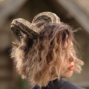 May include: A headpiece featuring two large, textured, curved ram horns in a metallic gold and black finish. The horns are attached to a band worn on the head, showcasing detailed ridges and a pointed tip on each side of the head.