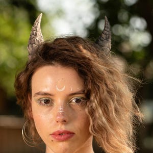 May include: A pair of dark gray, textured, curved horns attached to a headband. The horns feature a detailed, ridged surface pattern resembling natural bone or stone, positioned symmetrically on top of the head with a pointed, tapered shape.