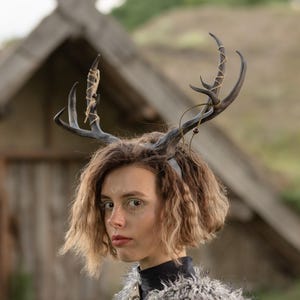 May include: A dark brown antler headband featuring realistic branching tines. The antlers are decorated with thin, light-coloured twine wrapped around the branches and small, dark spherical beads attached to the twine, secured on a simple black band.