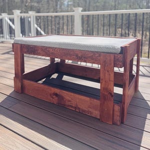 May include: A rectangular wooden bench with a dark brown stained finish and a light gray textured fabric cushion on top. The bench features a sturdy frame constructed from thick wooden planks with a lower shelf and four vertical corner supports.