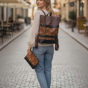 May include: A brown and black backpack with a snakeskin pattern on the front panel. The backpack has adjustable straps and a top handle. A matching clutch is also visible. The woman is wearing blue jeans and white shoes.