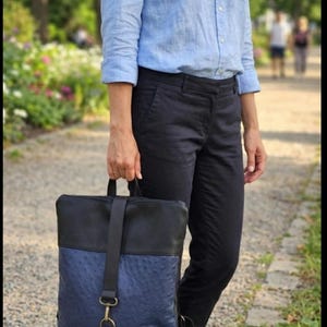 Pode incluir: Uma mochila preta e azul com um padrão texturizado. A mochila tem uma parte superior preta e uma frente azul texturizada com uma alça preta e ferragens douradas. A pessoa está vestindo uma camisa azul clara e calças pretas.