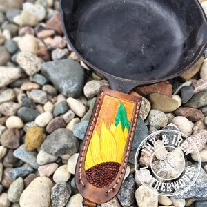 Op de afbeelding: Een bruine leren messenschede met een handgemaakt zonnebloemontwerp, gele bloemblaadjes en een bruin centrum. Naast de schede ligt een zwarte gietijzeren koekenpan. Het Ink & Iron Leatherworks-logo is zichtbaar.