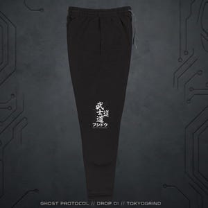Op de afbeelding: Zwarte joggingbroek met een trekkoord in de taille en witte Japanse tekst op het linkerbeen. De tekst luidt "Bushido: Way of the Warrior". De onderkant van de afbeelding heeft de tekst "GHOST PROTOCOL // DROP 01 // TOKYOGRIND".