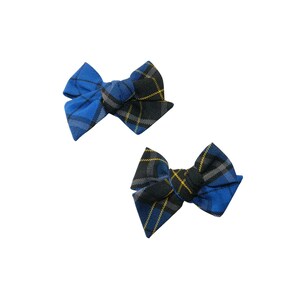 May include: Two blue and black plaid hair bows. The bows have a classic bow shape with a knot in the center. The plaid pattern includes blue, black, gray, and yellow lines. The bows are set against a white background.