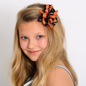 May include: A decorative hair bow featuring orange and black ribbons with white polka dots. The bow is attached to a hair clip and is worn on the side of the head. The bow is a festive accessory.