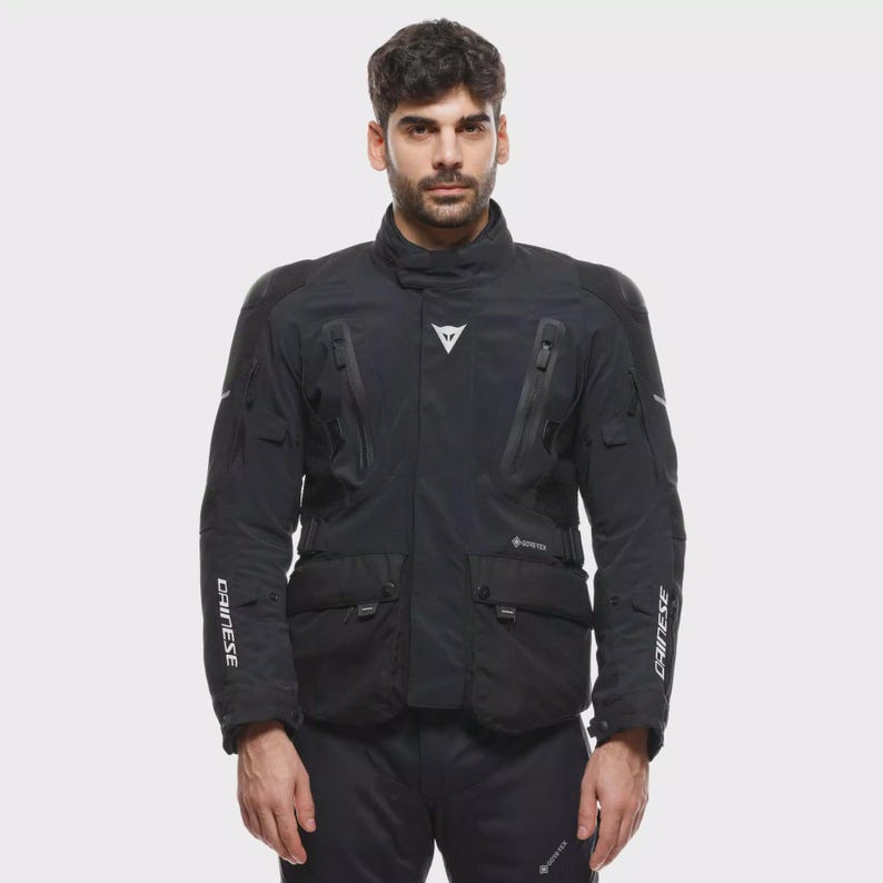 May include: Black motorcycle jacket with multiple pockets, zips, and the brand name "DAINESE" in white on the sleeves. The jacket has a high collar and a white logo on the chest. The jacket is made of a durable material and is designed for protection.