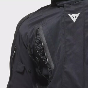 May include: Close-up of a black motorcycle jacket with a white logo. The jacket has several zipped pockets, one with a mesh lining. The image shows the details of the durable, weather-resistant fabric.
