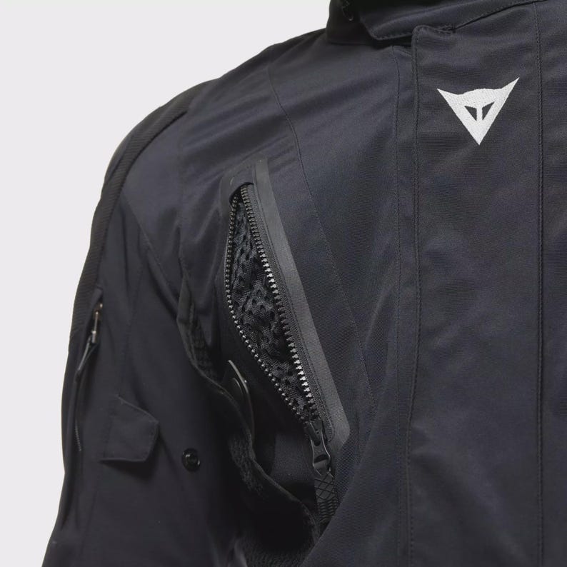 May include: Close-up of a black motorcycle jacket with a white logo. The jacket has several zipped pockets, one with a mesh lining. The image shows the details of the durable, weather-resistant fabric.