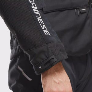 May include: Black motorcycle jacket sleeve with the white text "DAINESE" running vertically down the arm. The jacket has a textured fabric and a velcro cuff closure. The jacket is part of a motorcycle riding suit.