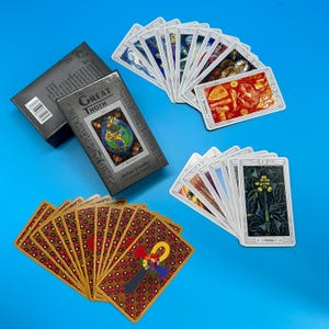 Könnte beinhalten: Ein Kartenspiel des Great Thoth Tarot mit einer Schachtel, auf einer blauen Oberfläche aufgefächert. Die Schachtel ist grau mit goldfarbenen Buchstaben und einem farbenfrohen Design. Die Karten haben komplizierte Illustrationen und ein goldenes Ankh-Symbol auf der Rückseite.