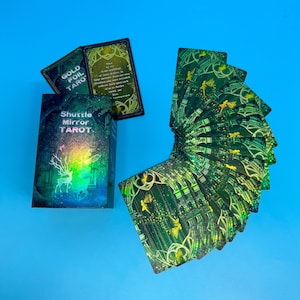 May include: A tarot card deck with a box that reads "Shuttle Mirror Tarot" and "Gold Foil Taro". The cards are green with gold accents and illustrations. The box features a deer illustration.