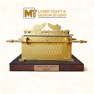 Puede incluir: Modelo dorado, cortado con láser, del Arca de la Alianza, con detalles intrincados y elementos decorativos. El modelo se asienta sobre una base marrón oscuro con una pequeña placa. La parte superior del arca tiene dos alas doradas. El texto "ARCA DEL PACTO" es visible.