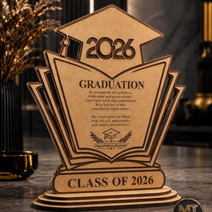 Puede incluir: Un premio de graduación de madera cortado con láser que presenta un birrete en la parte superior con el año 2026. El centro muestra una forma de libro abierto con el texto Graduation, un mensaje de felicitación y un icono de birrete.