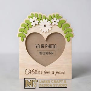 Puede incluir: Un marco de fotos de madera con un recorte en forma de corazón para una foto de 130 x 90 mm. El marco está decorado con hojas verdes y flores blancas. El texto "YOUR PHOTO" y "Mother's love is peace" son visibles.