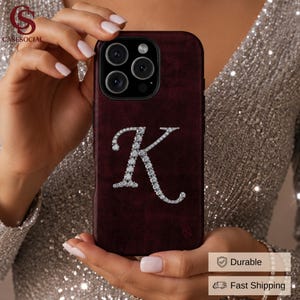 May include: A deep burgundy velvet-textured smartphone case featuring a large, centered letter K embellished with clear, sparkling crystals. The case has a cutout for a triple-lens camera system and a small logo in the bottom right corner.